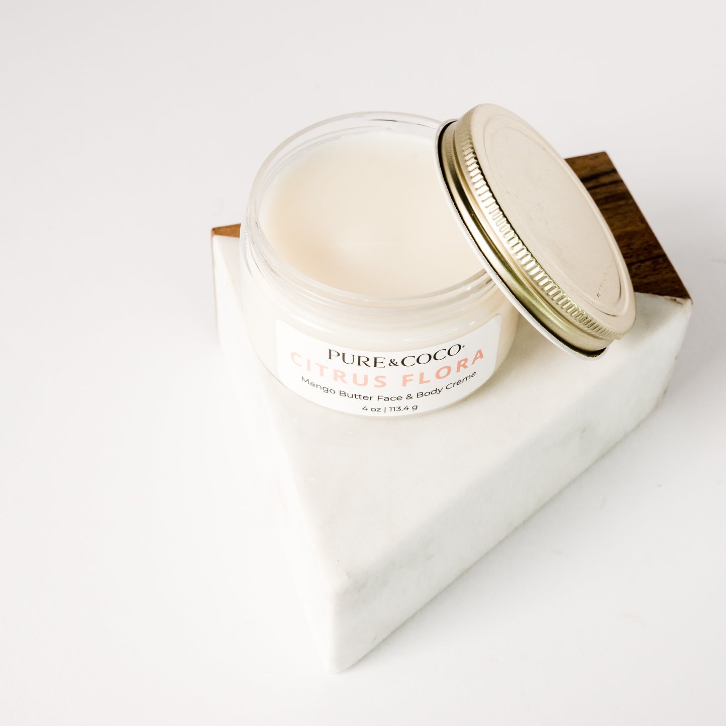Overlooking an open jar of the Citrus Flora Mango Butter Face & Body Creme_4 oz_Pure and Coco with the gold lid sitting at an angle next to the jar