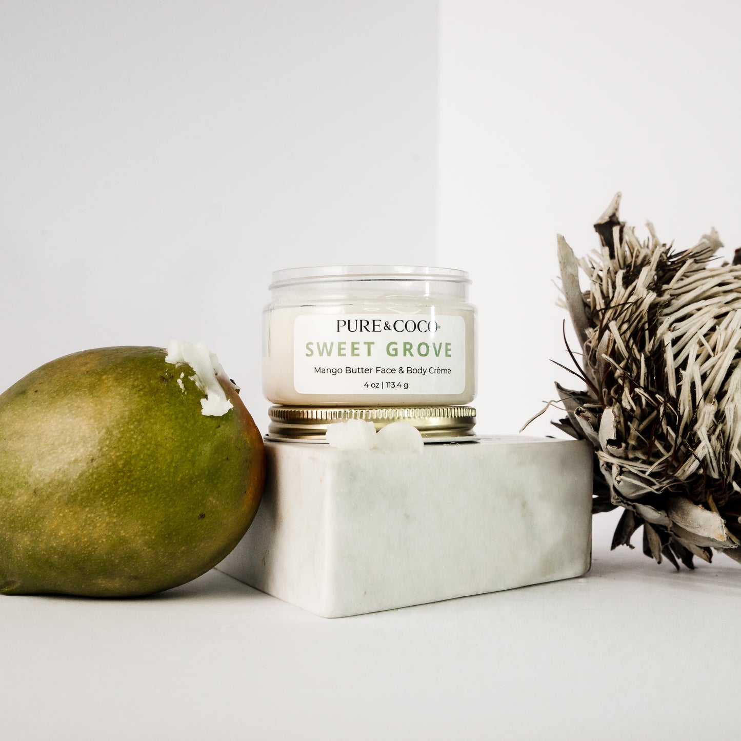 Flatlay of Sweet Grove Mango Butter Face & Body Crème surrounded by vanilla, sandalwood, and ylang ylang elements to highlight its natural scent profile.