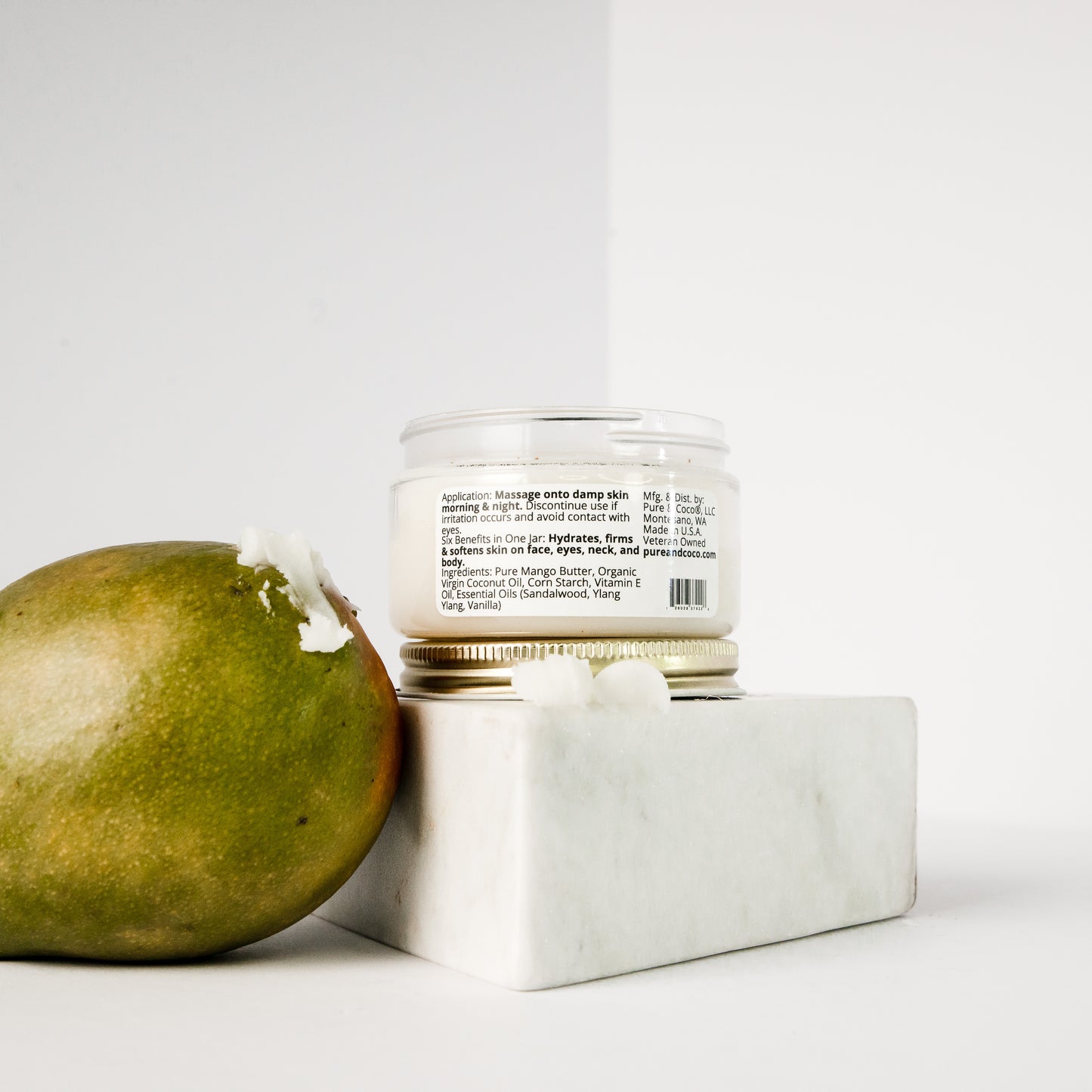 Sweet Grove Mango Butter Face & Body Crème jar displayed on a white marble block beside a fresh mango, representing the natural ingredients and purity of the formula.