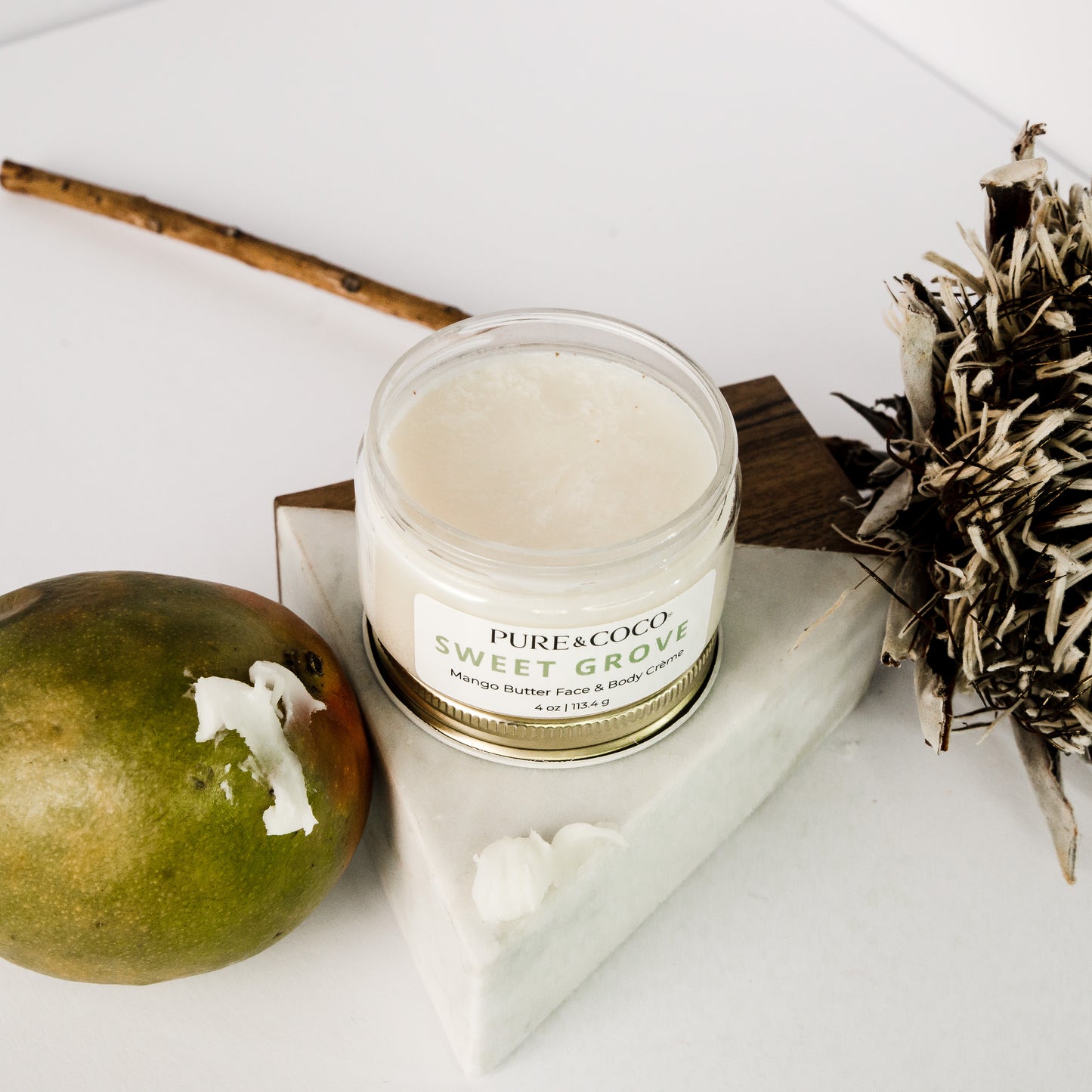 Mango sitting next to a jar of Sweet Grove Mango Butter Face & Body Creme_4 oz_Pure and Coco on a white background next to a flower