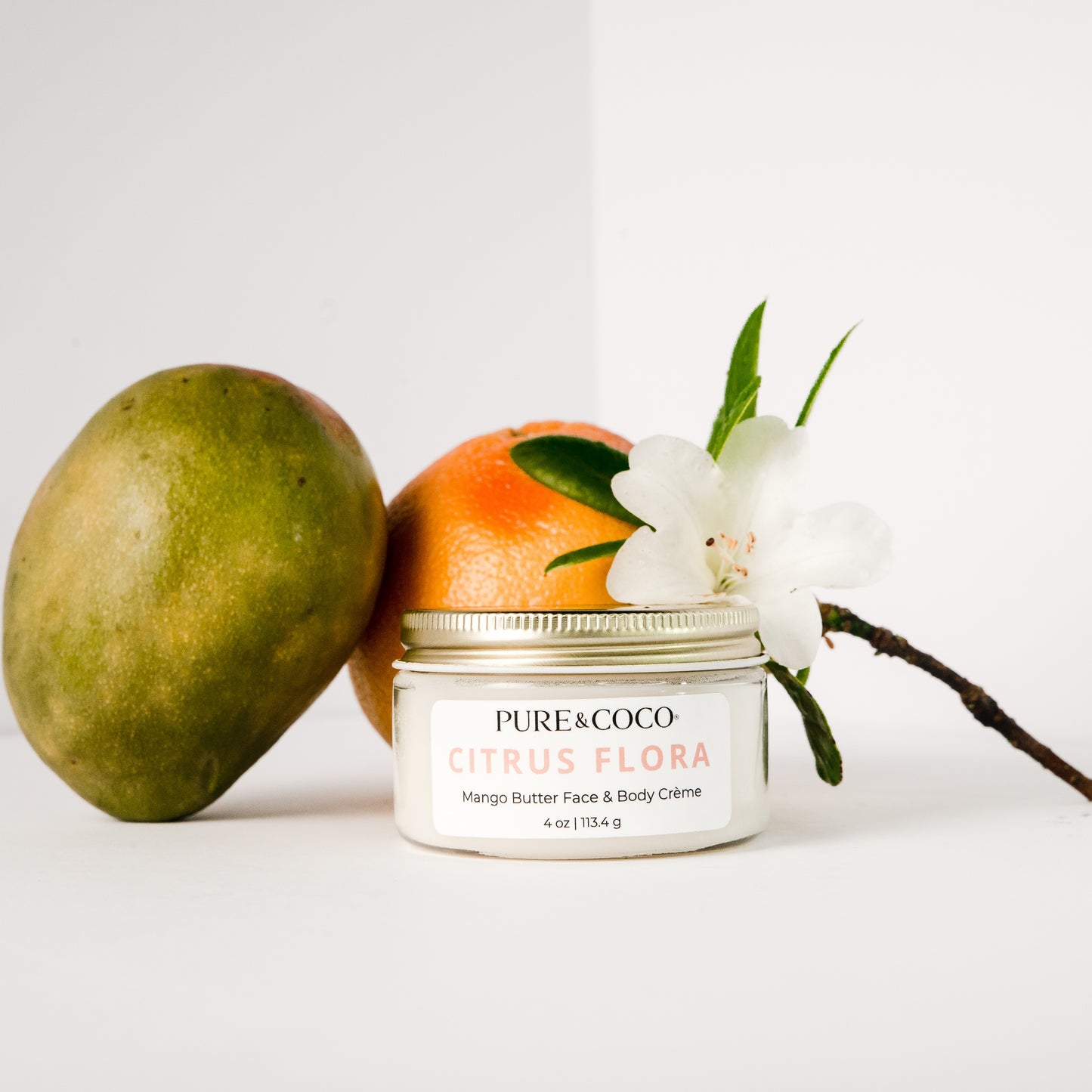 mango sitting next to the Citrus Flora Mango Butter Face & Body Creme_4 oz_Pure and Coco with a grapefruit and a flower surrounding it
