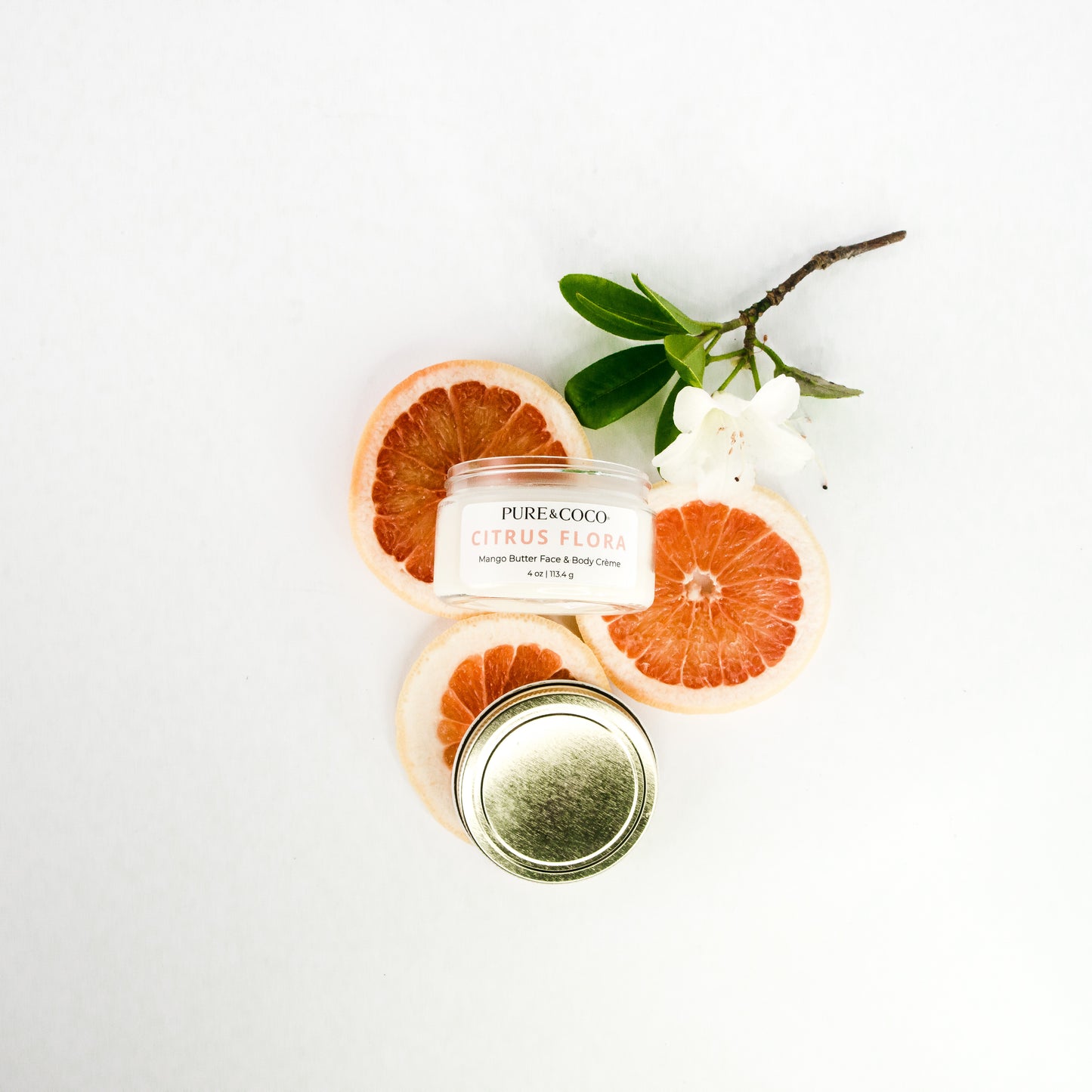 Flatylay of the Citrus Flora Mango Butter Face & Body Creme_4 oz_Pure and Coco on a white background with fresh slices of grapefruits and a flower to represent the scent