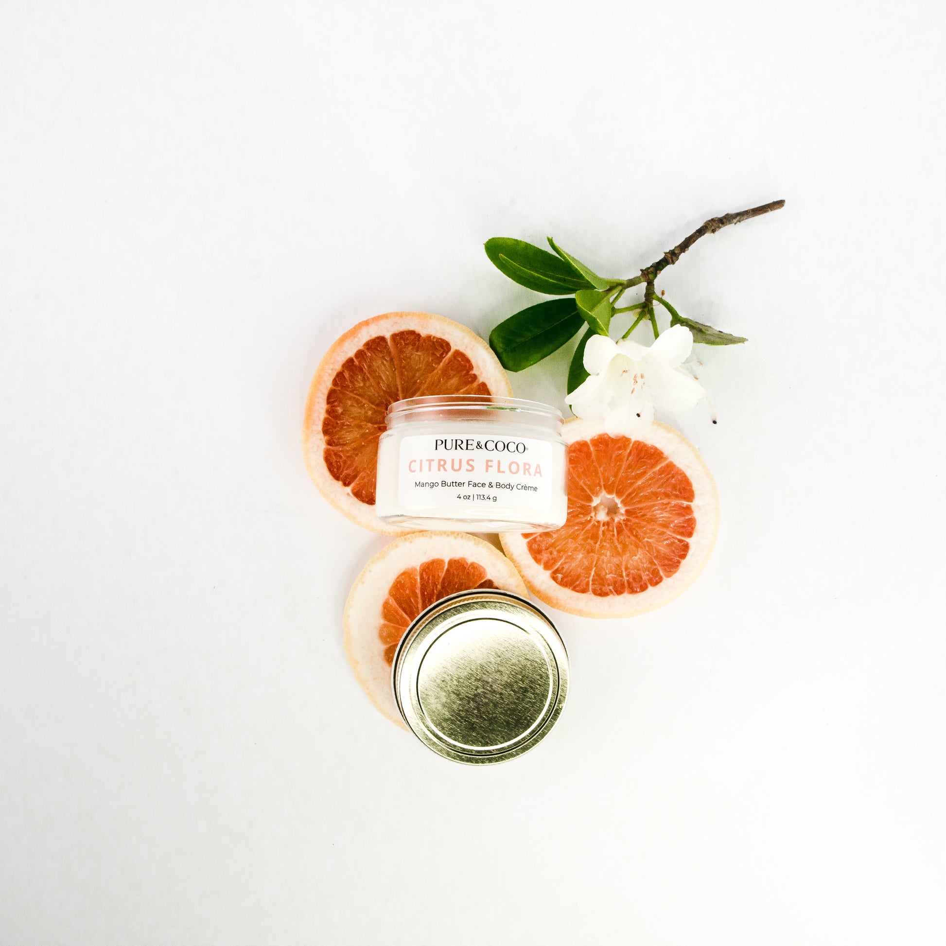 Flatylay of the Citrus Flora Mango Butter Face & Body Creme_4 oz_Pure and Coco on a white background with fresh slices of grapefruits and a flower to represent the scent
