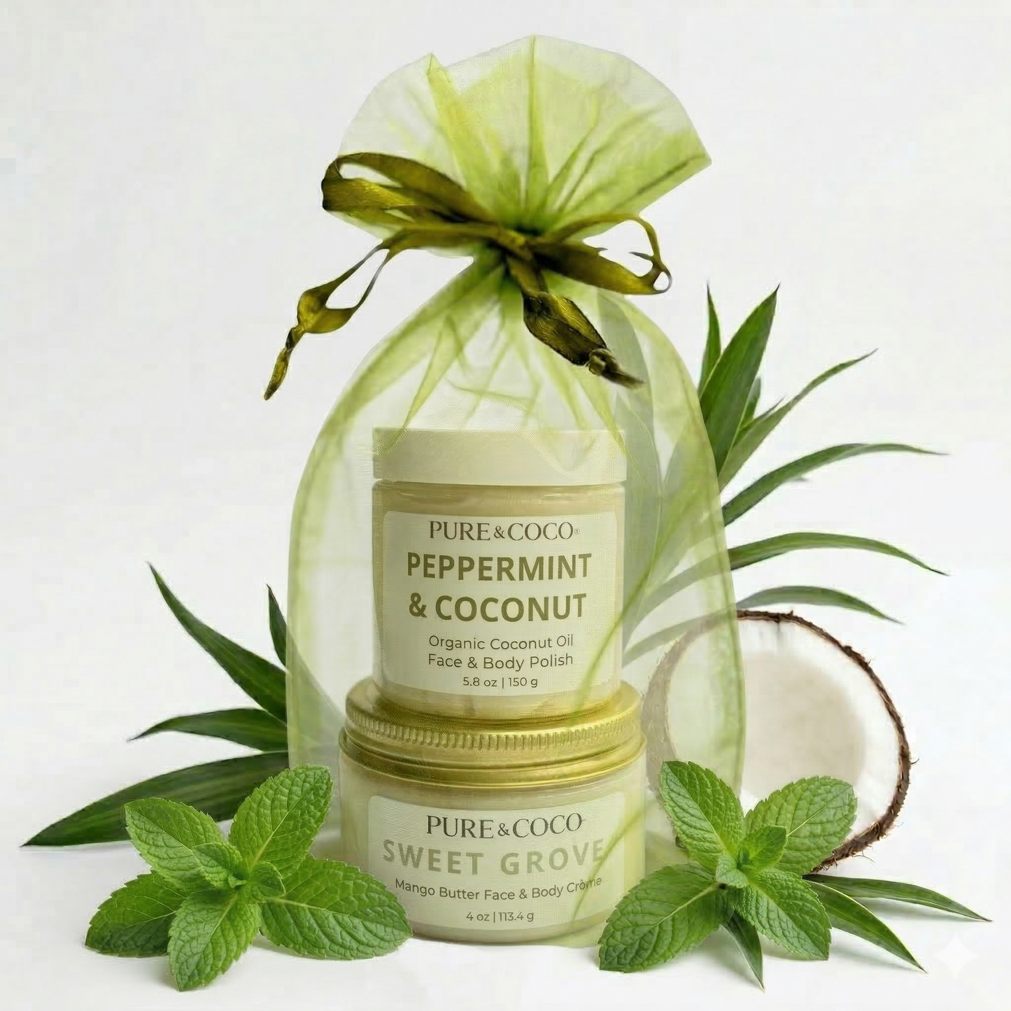 Pure & Coco Peppermint & Coconut Organic Body Polish and Mango Butter Crème Gift Set. Stacked jars are enclosed in a light green organza bag with a dark green ribbon, surrounded by fresh peppermint sprigs and a halved coconut, emphasizing organic, refreshing, and high-quality ingredients. Buy now for a premium, clean beauty experience.